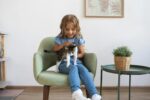 pexels-photo-5264047-5264047 A young girl sits on an armchair, gently holding a kitten beside a potted plant in a cozy room.